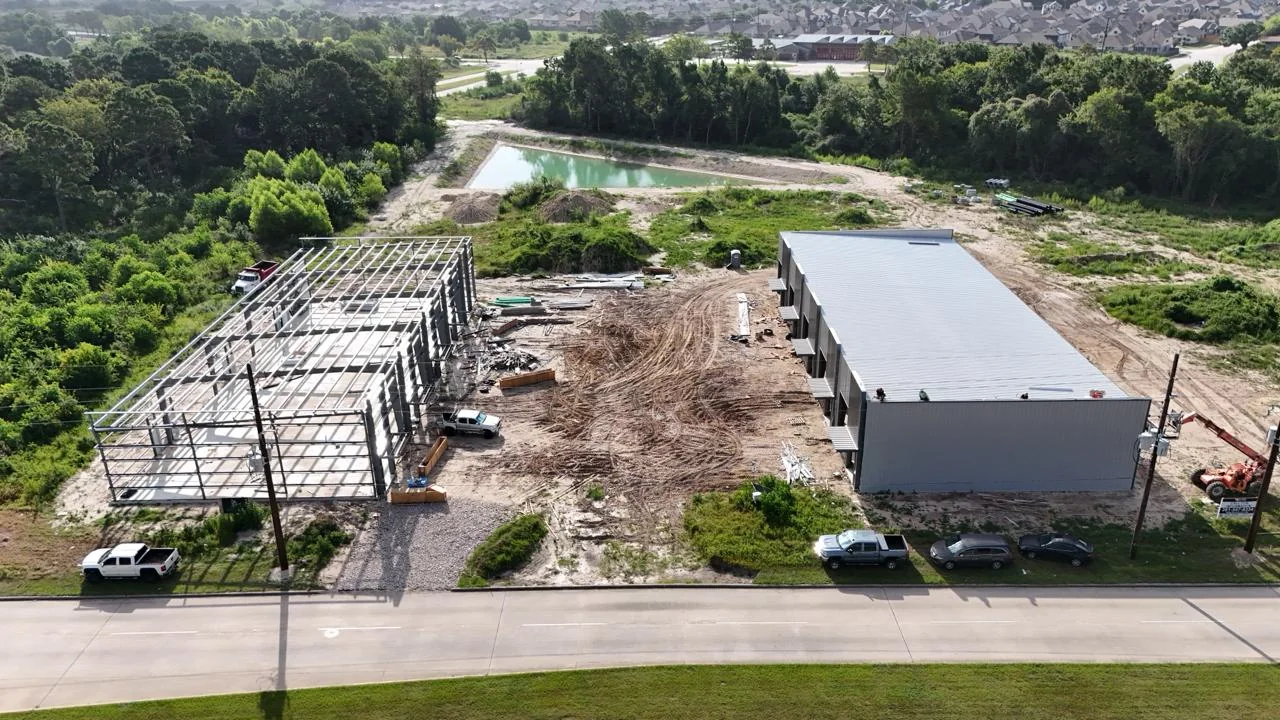 Day 166 aerial view: metal roof and exterior wall cladding installed on one building while structural steel framing continues on the adjacent building, August 14, 2026